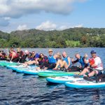 A large group pf paddleboarders rafted together in a long line on Coniston Water